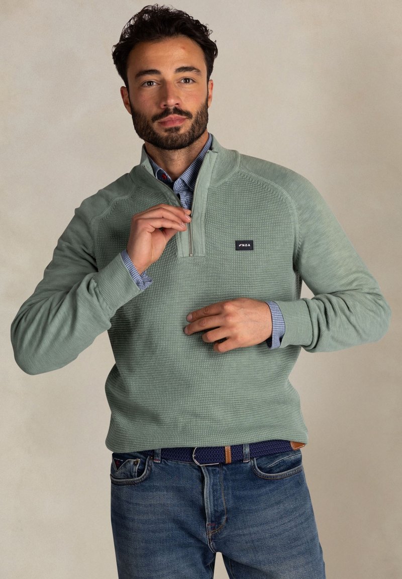 Lichtgroene kwart-rits pullover van textuurstof. Beschikt over een klein logopatch, geribde boorden en een casual pasvorm. Draagt over een gemusterd shirt.