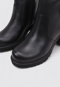 Botas de cuero negro hasta el tobillo con un acabado suave, punteras redondeadas, tacones elevados y suelas de goma texturizadas para mayor agarre. Cierre con cremallera lateral visible.