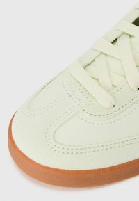 Lys grønn semsket sneaker med snøring, teksturerte overflatedetaljer og brun gummiksole. Rund tå og broderte detaljer.