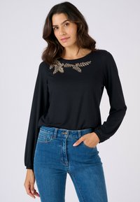 Top nero a maniche lunghe con ricamo decorativo a foglia dorata lungo la scollatura, abbinato a jeans di denim blu.