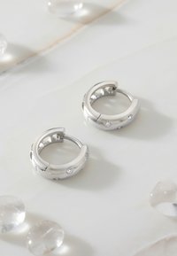 Silberne Creolen mit kleinen klaren Kristallen. Glattes, rundes Design mit Ausschnitten, auf einer marmorähnlichen Oberfläche ruhend.