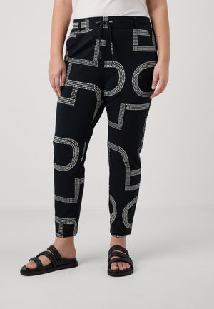 Schwarze Hosen mit weißen geometrischen Mustern, Tunnelzug in der Taille, kombiniert mit schwarzen Slide-Sandalen und einem weißen ärmellosen Top.
