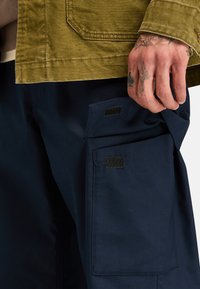 Olijfgroene jas met een gestructureerde afwerking, gecombineerd met marineblauwe cargobroeken met een grote zijzak en klittenbandaccenten.