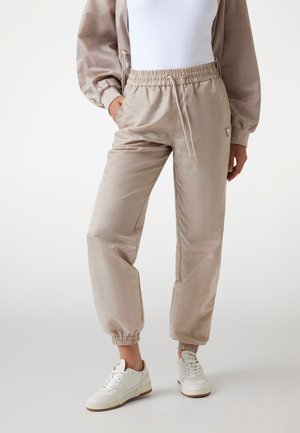 Beige jogger pants in a glossy fabric, featuring an elastic waistband with drawstring, ribbed cuffs, and subtle patterned detailing.