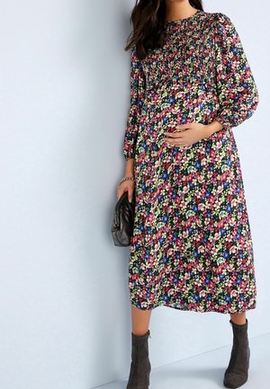 Vestido de maternidad floral en tela oscura con patrones coloridos, mangas largas, cuerpo fruncido y falda hasta el tobillo; combinado con botas grises.