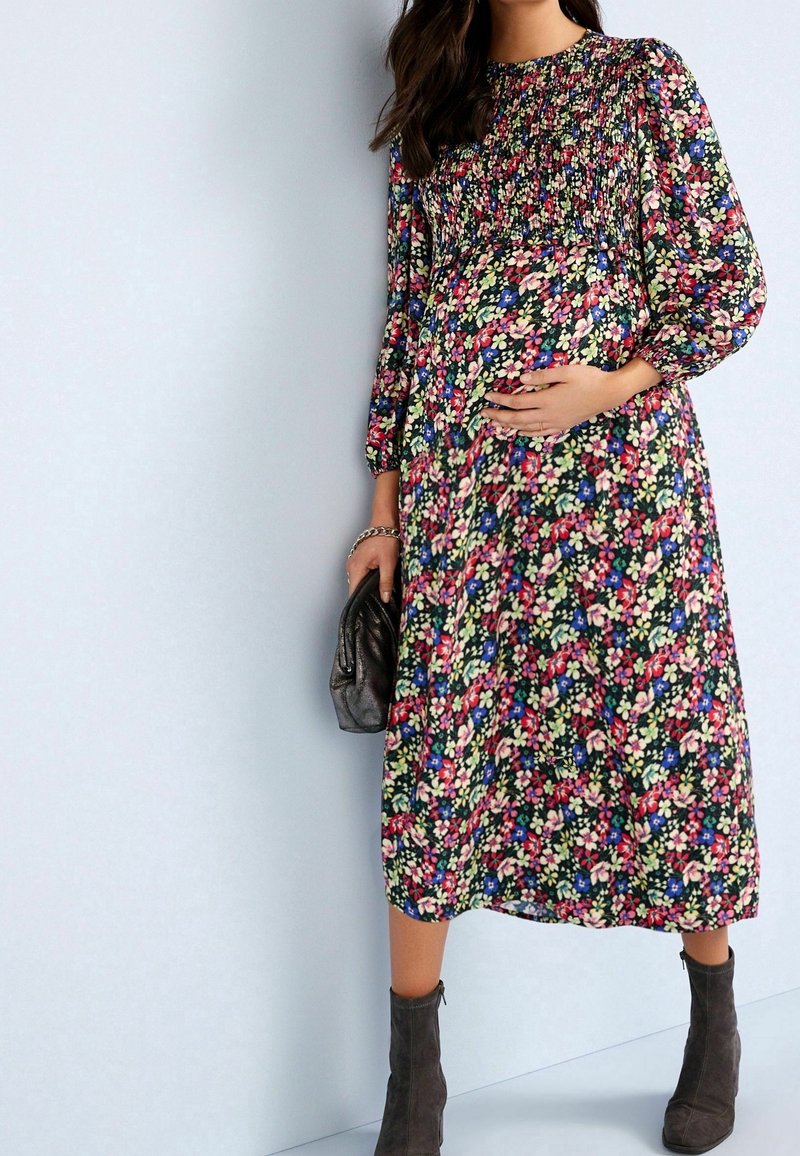 Vestido de maternidad floral en tela oscura con patrones coloridos, mangas largas, cuerpo fruncido y falda hasta el tobillo; combinado con botas grises.