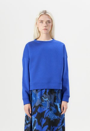 Blauer Rundhals-Sweatshirt aus weichem Material, mit überschnittenen Schultern und cropped Schnitt, kombiniert mit einem fließenden Rock im blauen Muster.