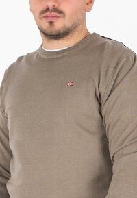 Maglione lavorato a maglia beige con collo rotondo e maniche lunghe. Distintivo emblema rosso e bianco sul petto. Tessuto liscio e testurizzato.