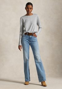 Kvinde iført lys grå langærmet skjorte, lyseblå jeans med høj talje, brunt bælte og lysebrune ankelstøvler på beige baggrund.