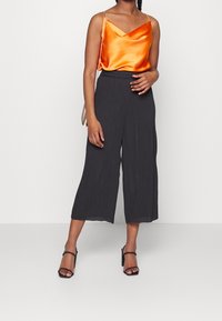 Eine Frau trägt ein ärmelloses Top aus orangefarbenem Satin, schwarze weite, knöchellange Hosen, schwarze Absatzsandalen und eine metallene Kettenhandtasche.