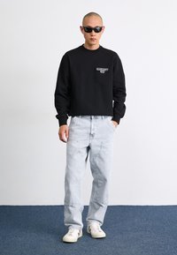 Black crewneck sweatshirt with front logo, light grey baggy jeans, and white sneakers with dark accents. Model wears black sunglasses.