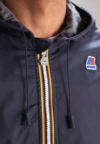 Giacca a vento blu navy con zip argentata, caratterizzata da strisce colorate e una fodera in tessuto. Logo sul petto sinistro, che evidenzia un design sportivo.