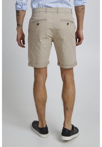 Pantalones cortos de algodón beige con bajo enrollado, dos bolsillos traseros con botones, llevados con zapatillas negras. Muestran una textura suave y un ajuste a medida.