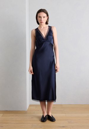 Kobieta stoi na drewnianej podłodze, ubrana w granatową, bezrękawnikową sukienkę midi z koronkowymi wykończeniami oraz czarne buty wsuwane.