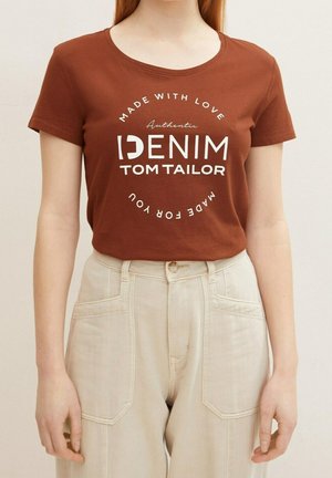 Kobieta w brązowej koszulce z krótkim rękawem z białym napisem "Authentic Denim Tom Tailor" i beżowych spodniach z wysokim stanem z przednimi kieszeniami.