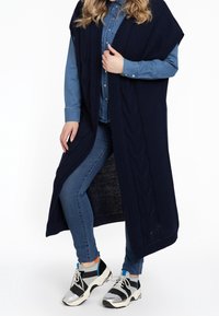 Granatowa dziergana kamizelka z wzorem warkoczy, noszona na dżinsowej koszuli. Do tego niebieskie dżinsy i czarne sneakersy z srebrnymi akcentami.