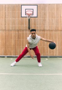 Ein junger Mann in einem grauen ärmellosen Hoodie und roten Jogginghosen dribbelt einen schwarzen Basketball auf einem grünen Platz mit einem Holzzaun im Hintergrund.