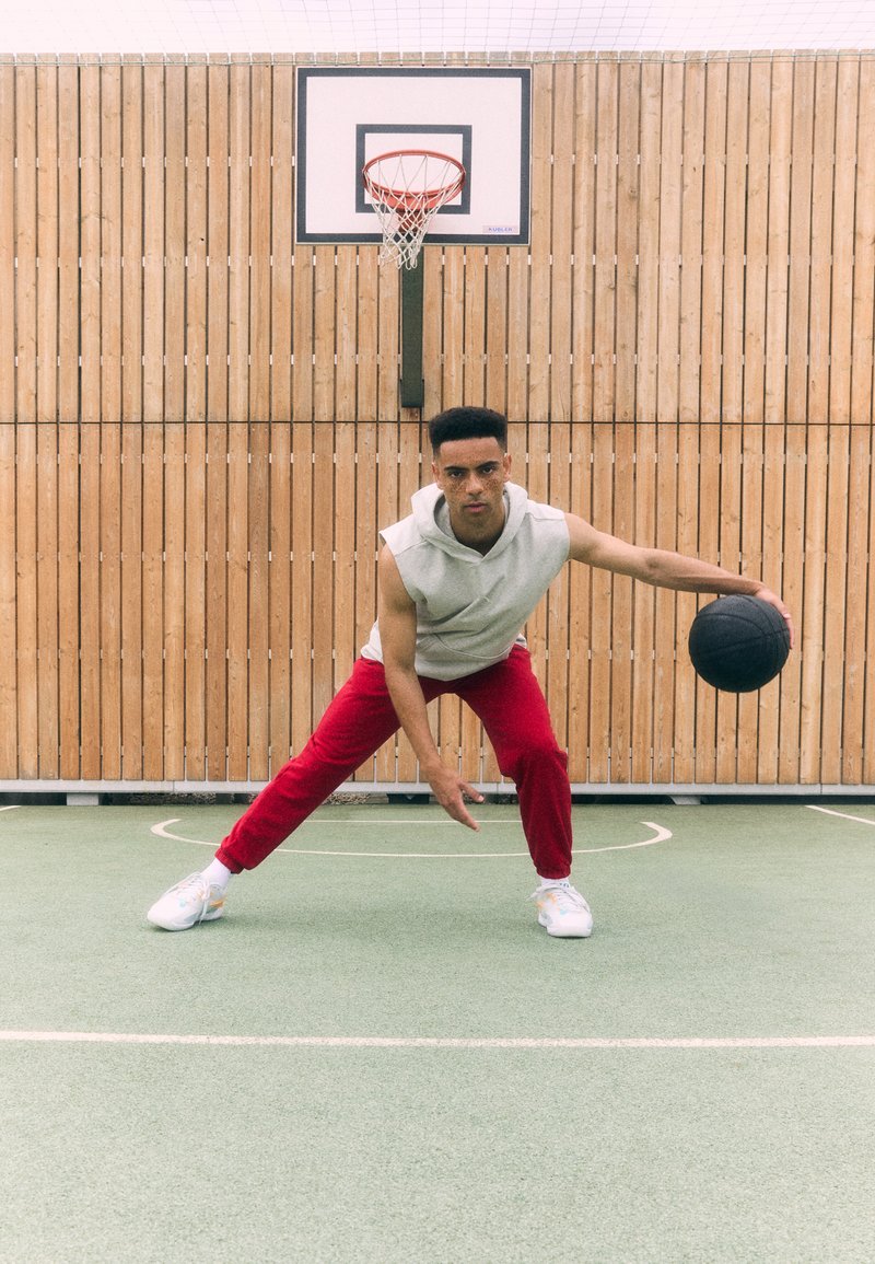 Un joven con un hoodie sin mangas gris y pantalones rojos dribla un balón de baloncesto negro en una cancha verde con un fondo de valla de madera.