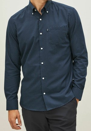 Homme portant une chemise bleu foncé à boutons avec un subtil petit motif carré et un pantalon foncé, main reposant négligemment dans la poche.