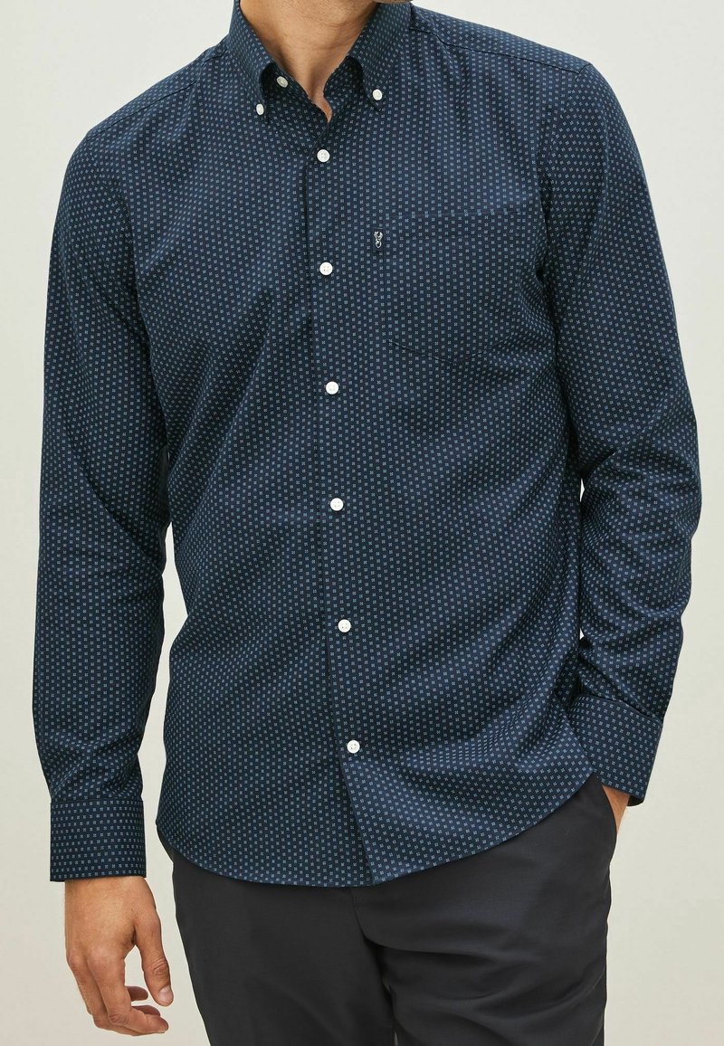 Homme portant une chemise bleu foncé à boutons avec un subtil petit motif carré et un pantalon foncé, main reposant négligemment dans la poche.