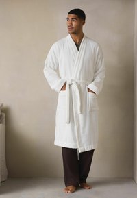 Weißer Frottee-Bademantel mit langen Ärmeln, Gürtel und Taschen, kombiniert mit braunen strukturierten Hosen. Barfuß, stehend gegen eine neutrale Wand.