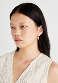 Gulltone øredobber med dråpeform, hengende fra en liten bøyle. Modellen har på seg en teksturert, lysbluse med V-hals.
