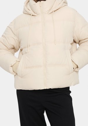 Béžová prošívaná péřová bunda s kapucí, předním zipem, šňůrkami a bočními kapsami, nošená s černými kalhotami. Ruce v kapsách.