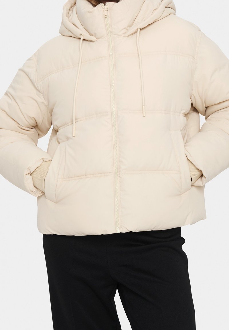 Veste matelassée beige avec capuche, fermeture éclair à l'avant, cordons de serrage et poches latérales portée avec un pantalon noir. Mains dans les poches.