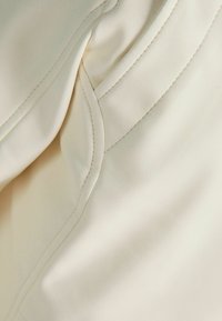 Kremowa tkanina o gładkiej fakturze, z zaokrąglonymi szwami i subtelnymi detalami przeszyć, nadająca ubraniu dopasowany wygląd.