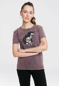 Grafisches T-Shirt aus verblasstem lila Stoff mit einem Cartoon-Design von einem Maulwurf. Kurze Ärmel, Rundhals-Ausschnitt und lockere Passform. Sichtbare schwarze Hose.