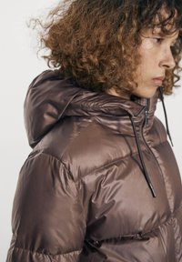 Profilbillede af en person med krøllet hår iført en brun dunjakke med hætte og sorte snore mod en ensfarvet baggrund.