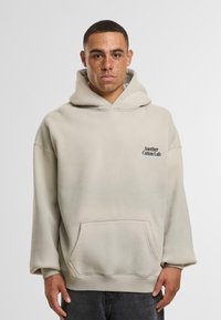 Uomo con lentiggini che indossa una felpa beige con tasca frontale e il logo "Another Cotton Lab" sul petto, in piedi davanti a uno sfondo semplice.