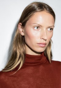 A close-up of a person with long, straight blonde hair, wearing a fitted rust-colored turtleneck top, against a white background.