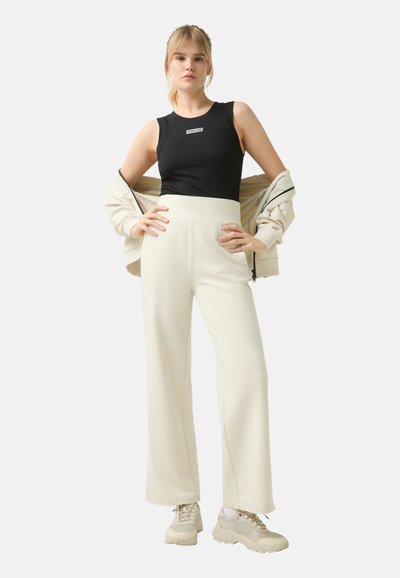 Black ribbed tank top with a logo, paired with cream wide-leg pants and beige athletic shoes. The model wears a light jacket.