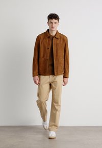 Brown suede jacket with front buttons and two large pockets, paired with beige trousers and white sneakers. Minimalist, clean design.