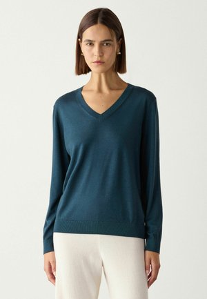Femme aux cheveux bruns raides portant des boucles d'oreilles dorées, un pull bleu sarcelle à col en V et un pantalon blanc sur un fond clair uni.