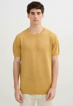 Joven con pelo rizado y oscuro que lleva un jersey texturizado de manga corta color mostaza y pantalones beige claros, de pie frente a un fondo liso.