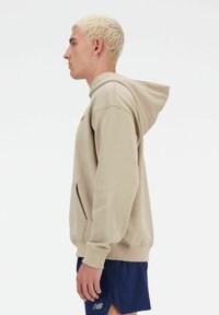 Felpa con cappuccio beige con tasca a canguro, realizzata in tessuto morbido. Presenta polsini e orlo a costine, cuciture in vista e un accento con il logo.