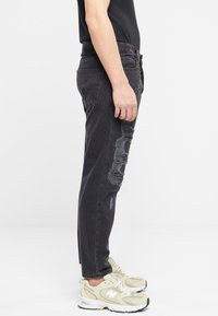 Vaqueros de mezclilla negra con detalles desgastados a lo largo de los lados, con un corte relajado y diseño de pierna recta. Llevados con zapatillas de color claro.
