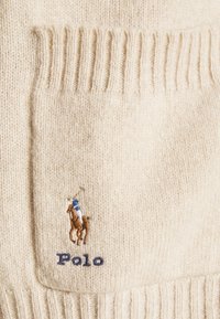 Beige pletený svetr s ribbed texturou. Obsahuje přední kapsu, vyšívané polo logo v hnědé a modré barvě a nápis "Polo" tmavě modrou.