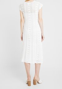Abito a maglia bianco con maniche corte a cappuccio, caratterizzato da un motivo testurizzato e una silhouette al ginocchio. Indossato con sandali bianchi con tacco.