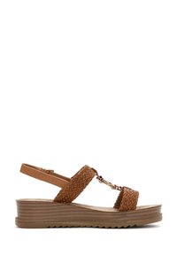 Sandalias trenzadas marrones con suela de cuña. Las características incluyen una correa de tobillo ajustable y herrajes metálicos decorativos en la parte delantera. Construcción ligera y texturizada.