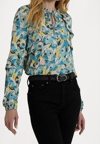 Barevná květinová blůza s modrými, žlutými a hnědými vzory. Má zavazování na krku a volánkové rukávy. Kombinováno s černými džíny.