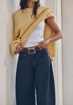 Žena podešava smeđi remen na hlačama širokih nogavica s prugama, nosi bijelu majicu bez rukava i žuti džemper prebačen preko ramena, ispred bež zida.