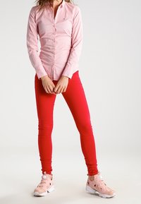 Lyserød skjorte med knapper og lange ærmer, kombineret med stramme røde bukser og lyserøde træningssko med hvide detaljer og et voluminøst design.