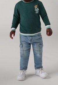 Grüner Sweatshirt mit weißen und mintfarbenen Rippb�ündchen, mit einem Bärendesign. Hellblaue Cargo-Jeans mit Taschen. Weiße Turnschuhe.