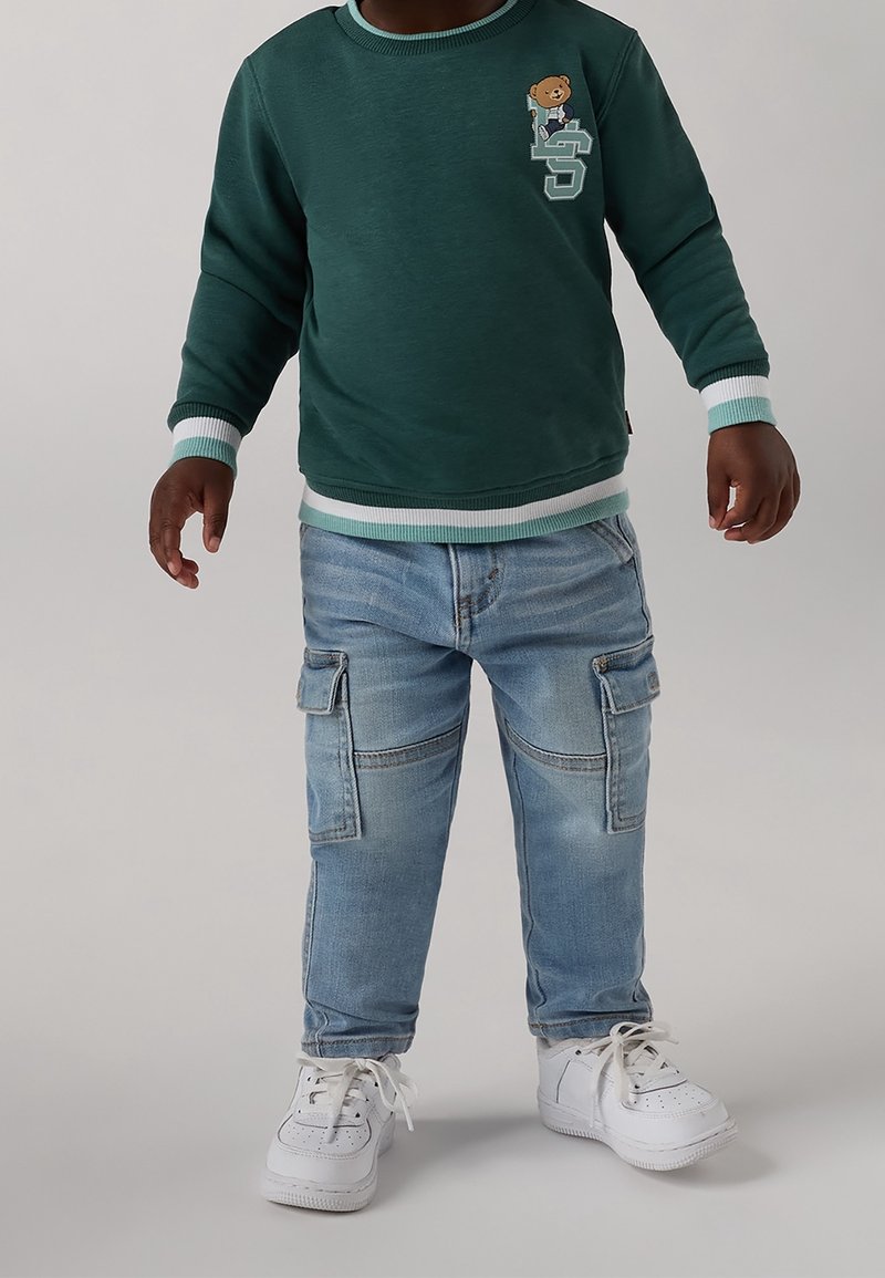 Grüner Sweatshirt mit weißen und mintfarbenen Rippbündchen, mit einem Bärendesign. Hellblaue Cargo-Jeans mit Taschen. Weiße Turnschuhe.
