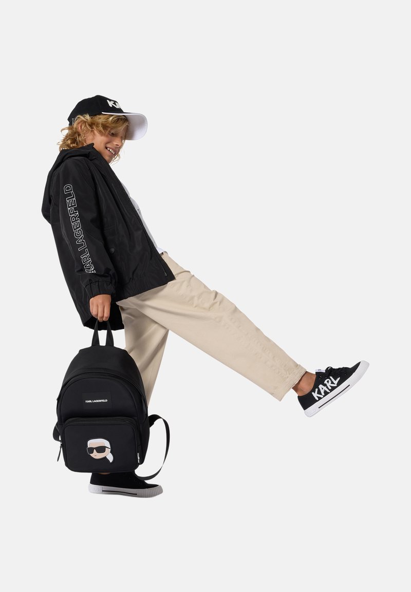 Sac à dos noir avec un graphique de personnage, associé à un coupe-vent noir, un chino beige et des baskets noires avec des accents blancs.