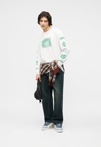 Νεαρό άτομο που φοράει λευκό μακρυμάνικο μπλουζάκι με γραφικό σχέδιο, σκούρα τζιν με φαρδιά μπατζάκια, αθλητικά παπούτσια, πουλόβερ καρό δεμένο γύρω από τη μέση και κρατάει μια μαύρη τσάντα.