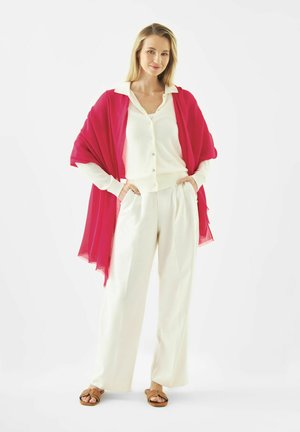 Femme debout, les mains dans les poches, portant une chemise blanche et un pantalon, un châle rouge et des sandales beiges, souriante sur un fond blanc.